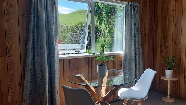 A room with a glass table and white chairs, a small wooden side table with a plant, and a window with curtains. -  Captivating rural beauty, serene getaway, nestled midway  Rotorua & Taupo. (Rerewhakaaitu)