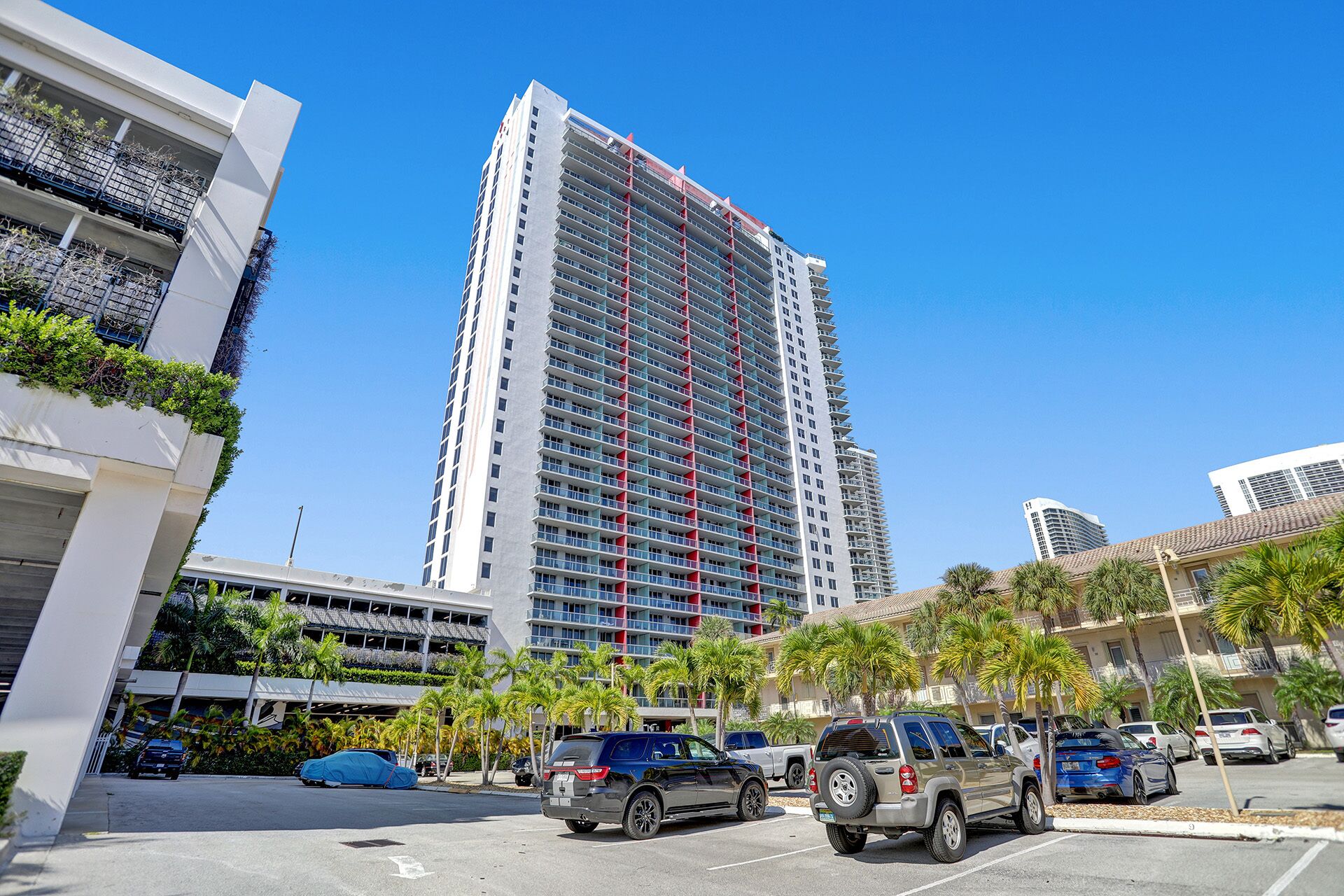 A tall hotel with multiple floors, a parking lot with several cars, and palm trees in the foreground.