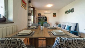 A dining room with a long wooden table set for four, surrounded by patterned chairs. There is a sofa with blue cushions against the wall, and a window with curtains on the left.