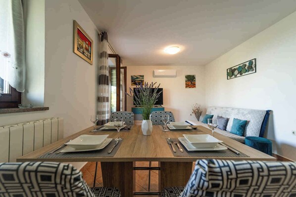 A dining room with a long wooden table set for four, surrounded by patterned chairs. There is a sofa with blue cushions against the wall, and a window with curtains on the left.