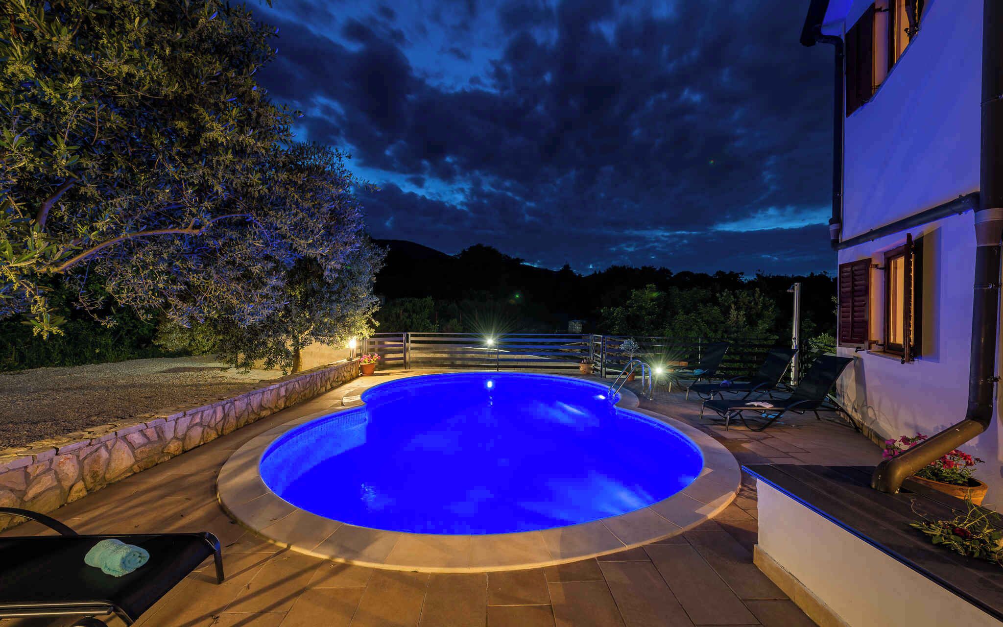A swimming pool with a blue-lit edge, surrounded by a stone wall and a wooden deck. There are lounge chairs and a potted plant nearby.