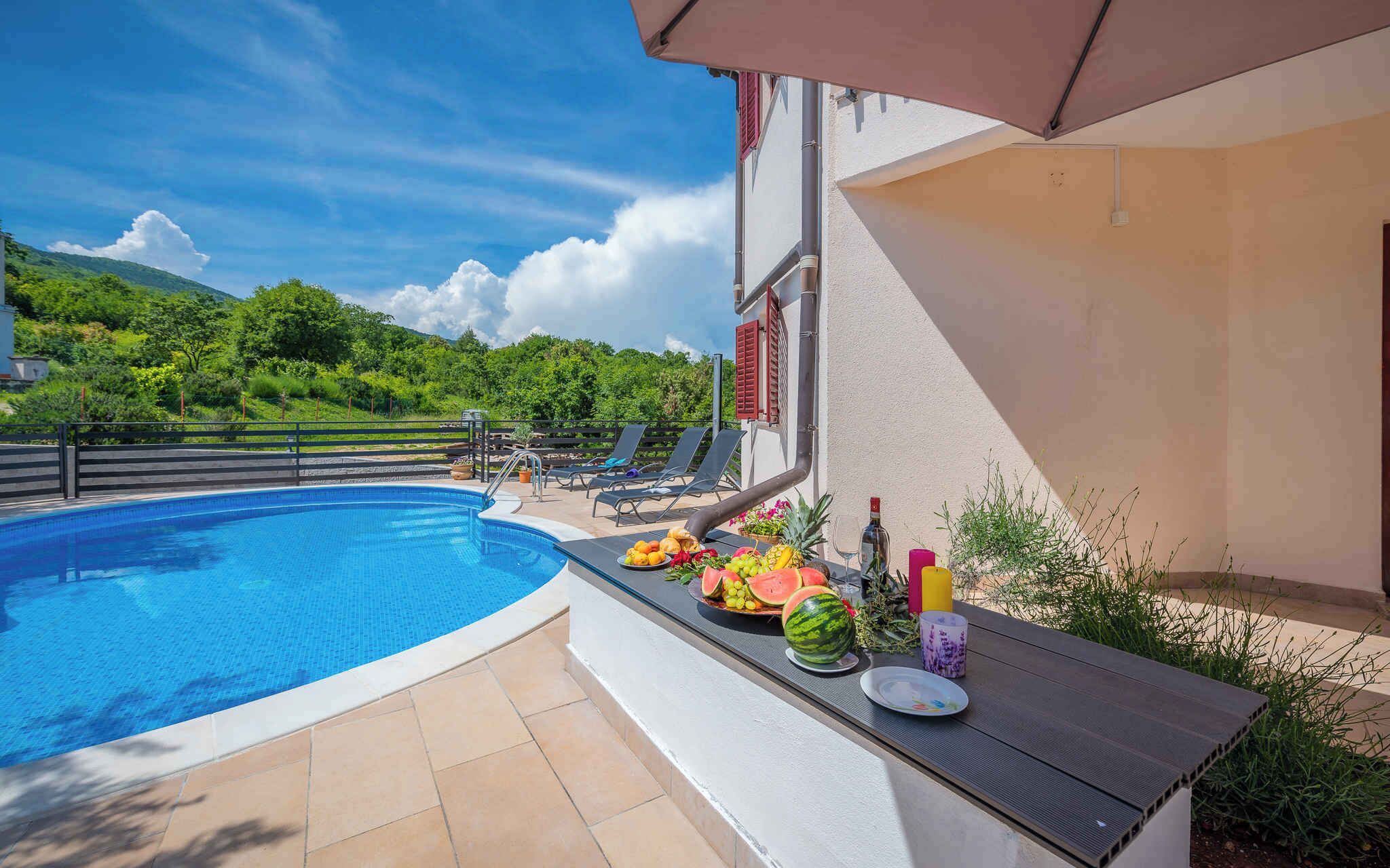 A swimming pool with a tiled deck, a table with fruits and drinks, and a view of a green landscape.