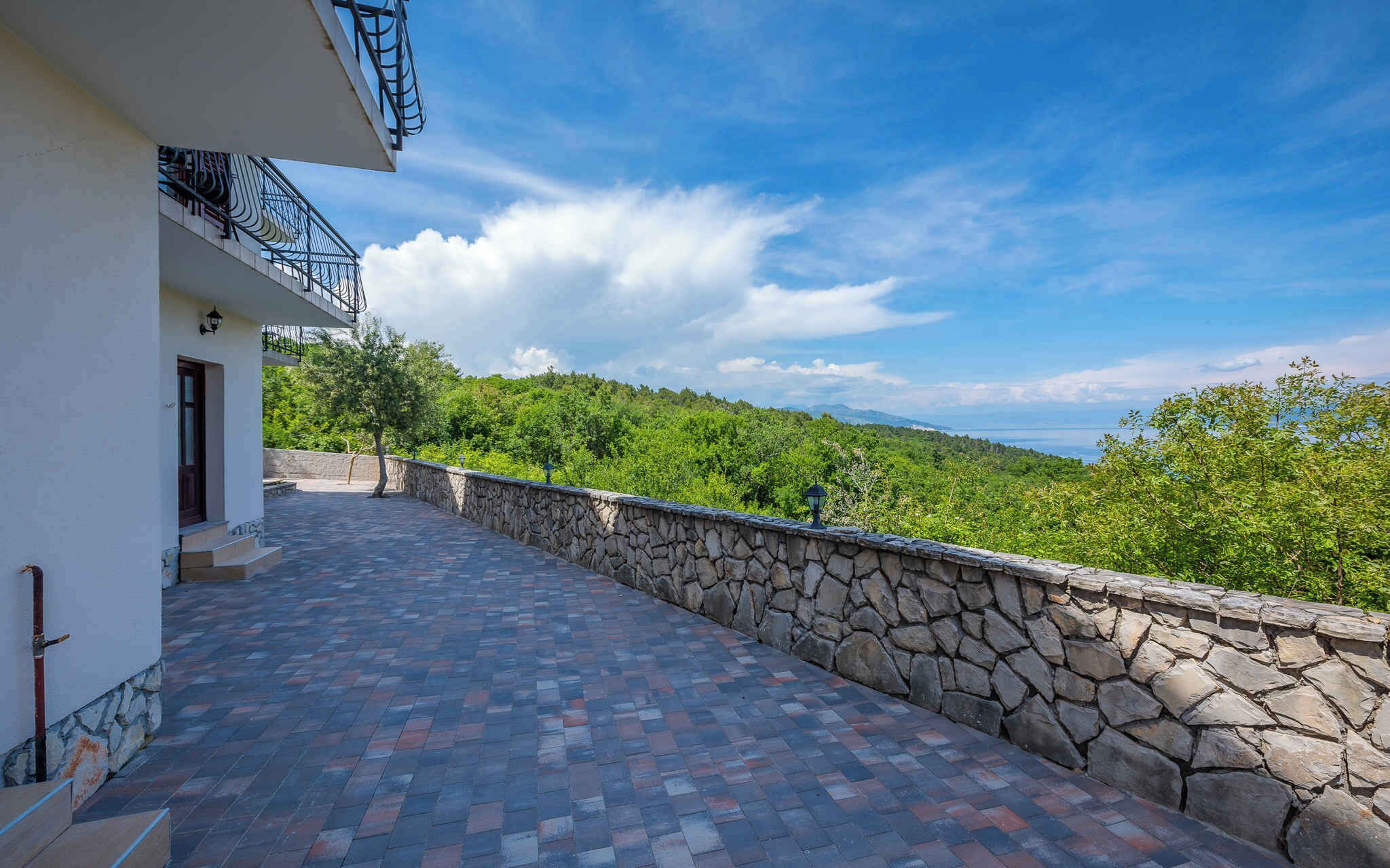 Ein Balkon mit Steinwänden und Fliesenboden, der einen üppigen, grünen Landschaftsbereich und ein Gewässer überblickt.