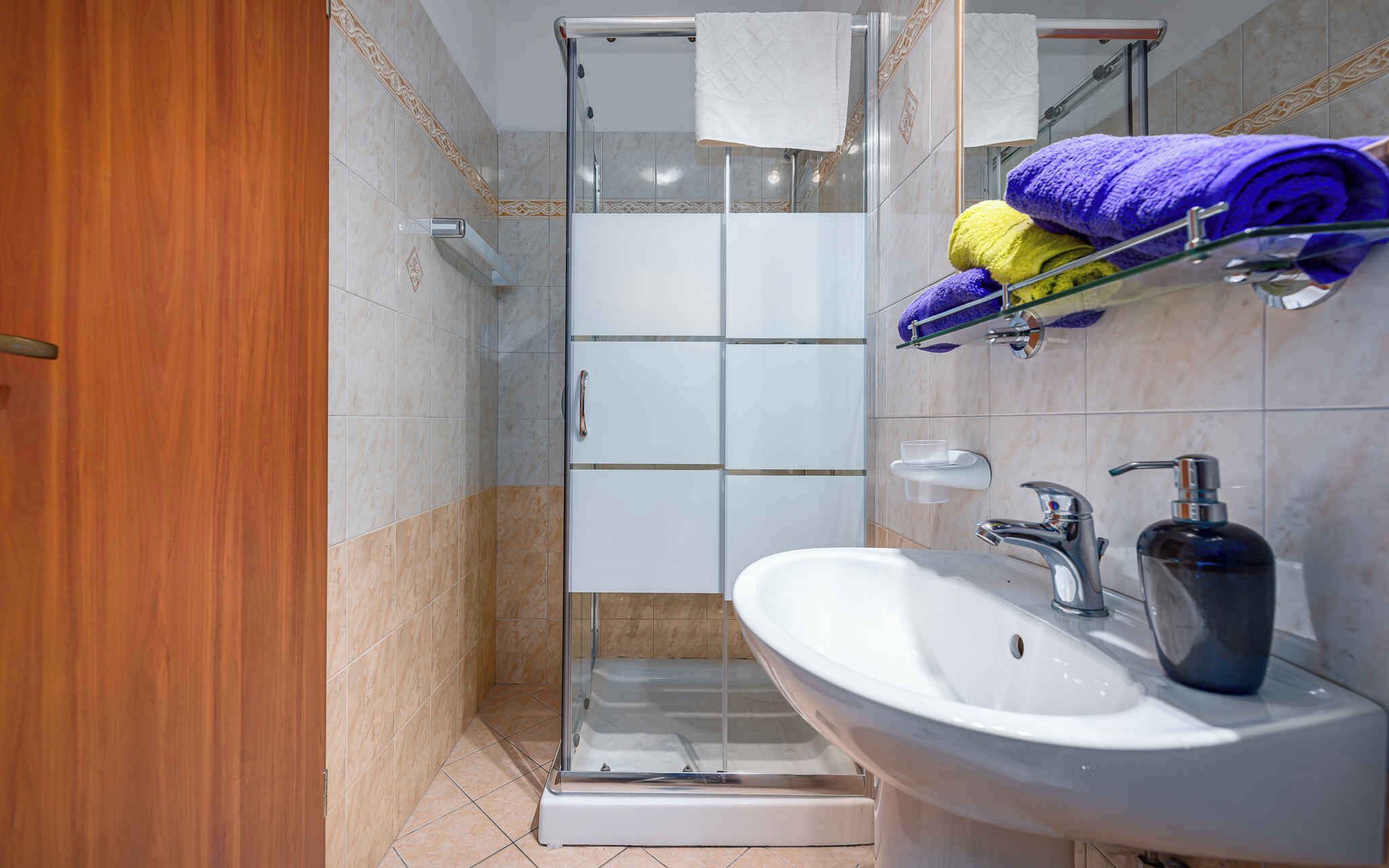Un baño moderno con cabina de ducha de vidrio, un lavabo blanco y un perchero con toallas moradas y amarillas.