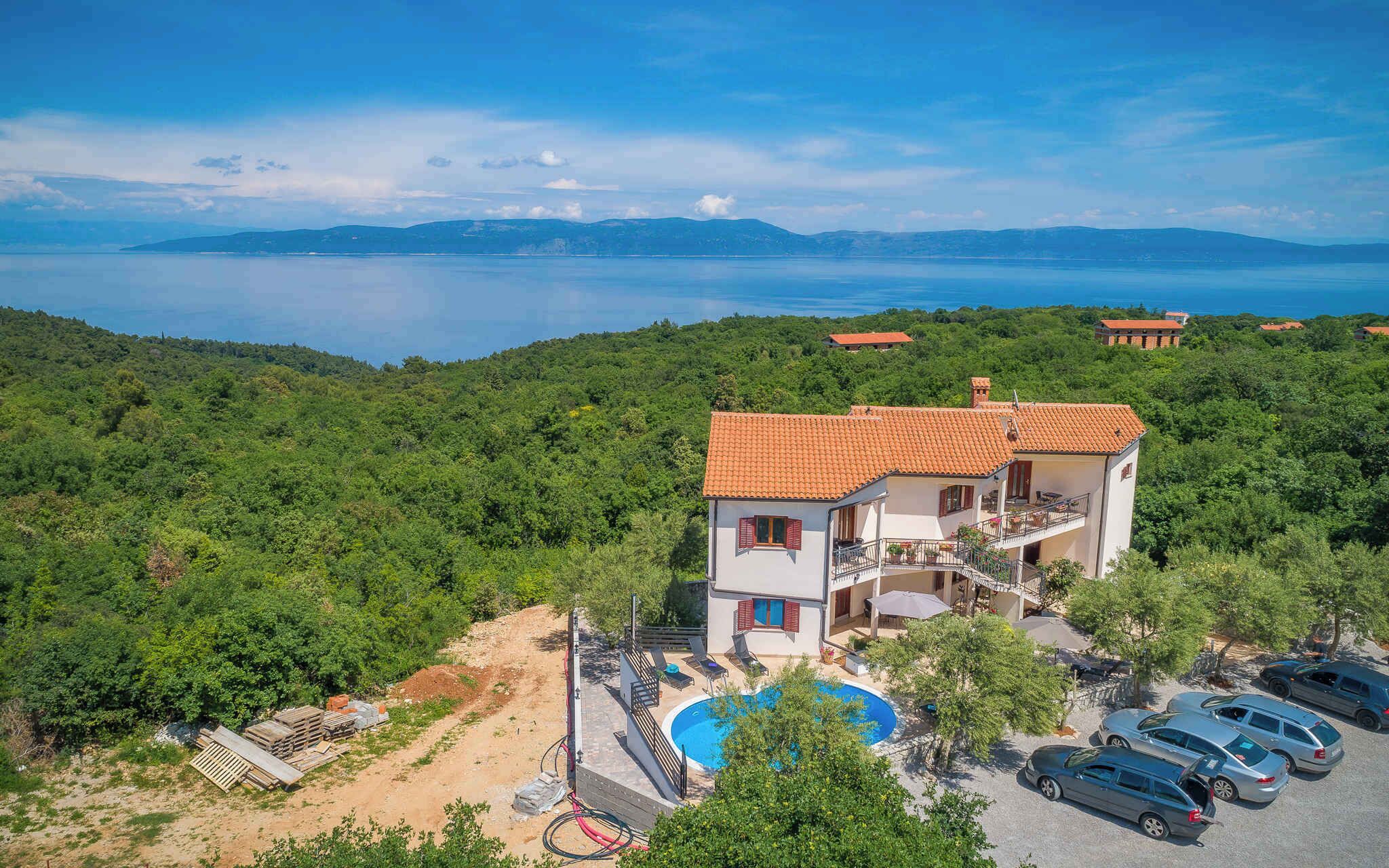 A two-story villa with a swimming pool, surrounded by lush greenery and a view of the sea.