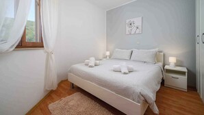 A bedroom with a bed, bedside tables, a nightstand, a window with sheer curtains, and a wall-mounted artwork.