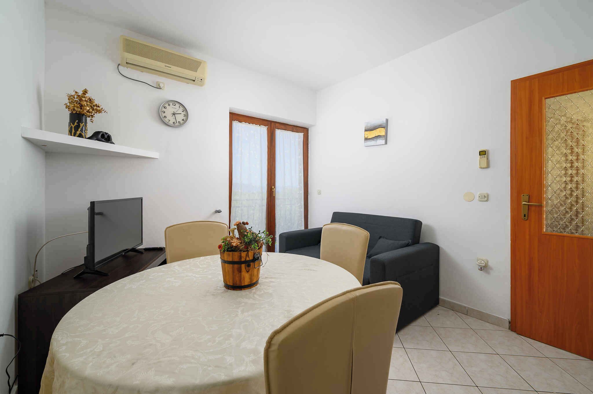 A small, well-lit room with a round dining table, chairs, a television, a sofa, a wooden door, and a wall-mounted air conditioner.