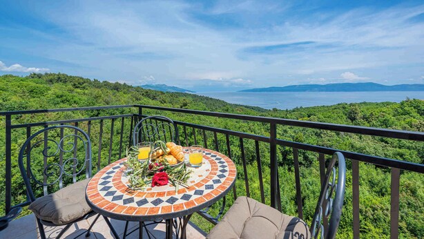 A round table with a mosaic tile design, surrounded by metal chairs, set on a balcony overlooking a lush green landscape and a body of water. - Holiday flat with air conditioning (Labin)