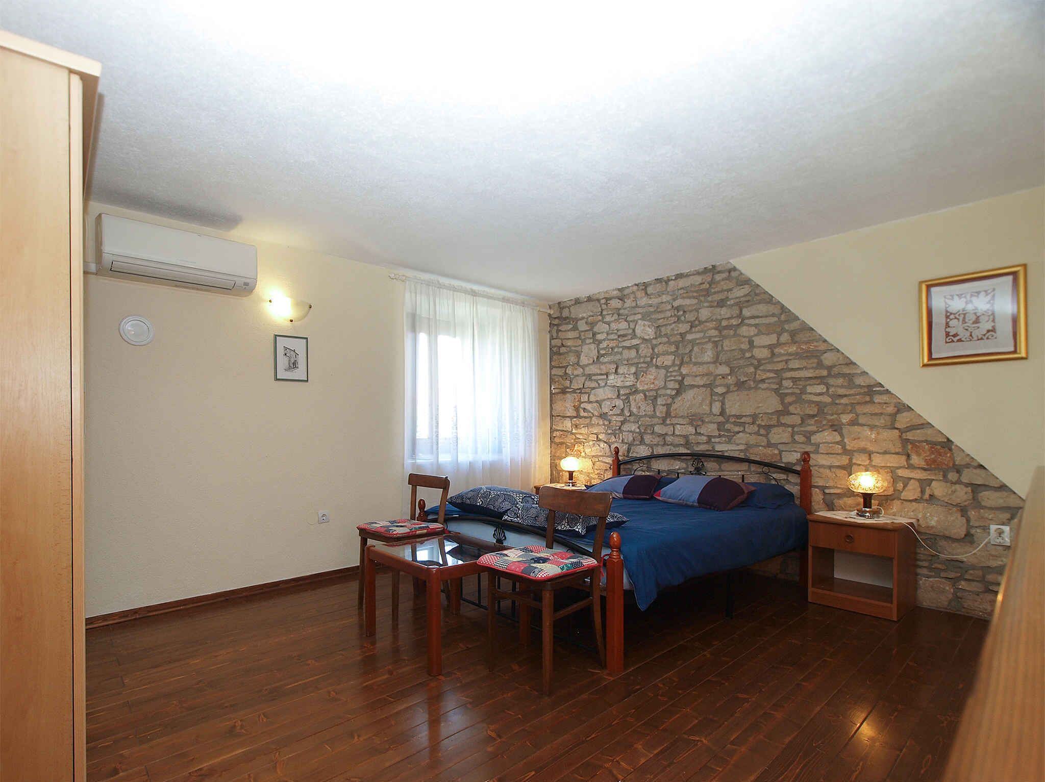 A bedroom with a stone wall, a bed with blue bedding, a wooden table with chairs, and a window with curtains.