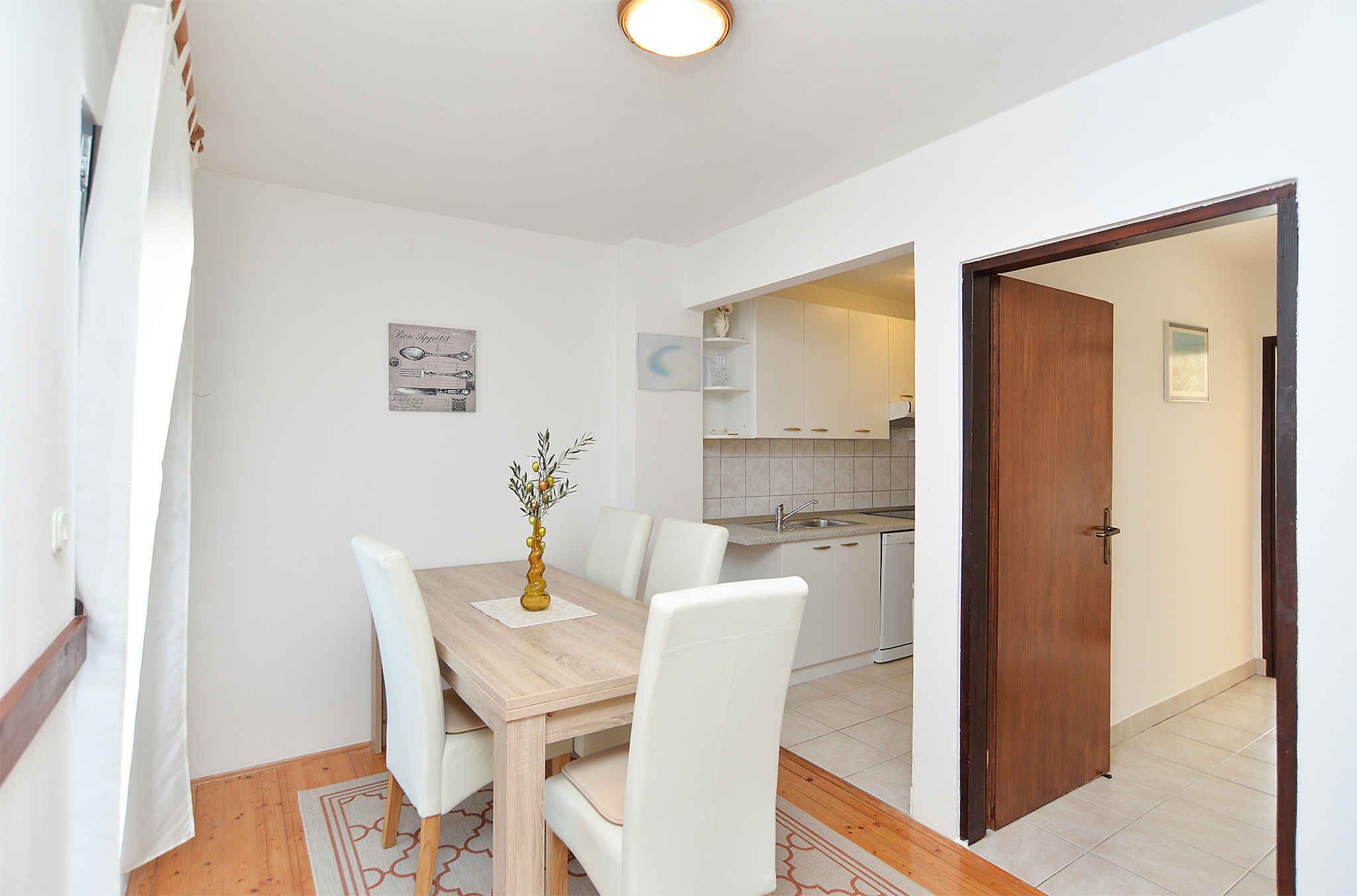 Een eethoek met een houten tafel en witte stoelen, een vaas met bloemen, en een keuken op de achtergrond.