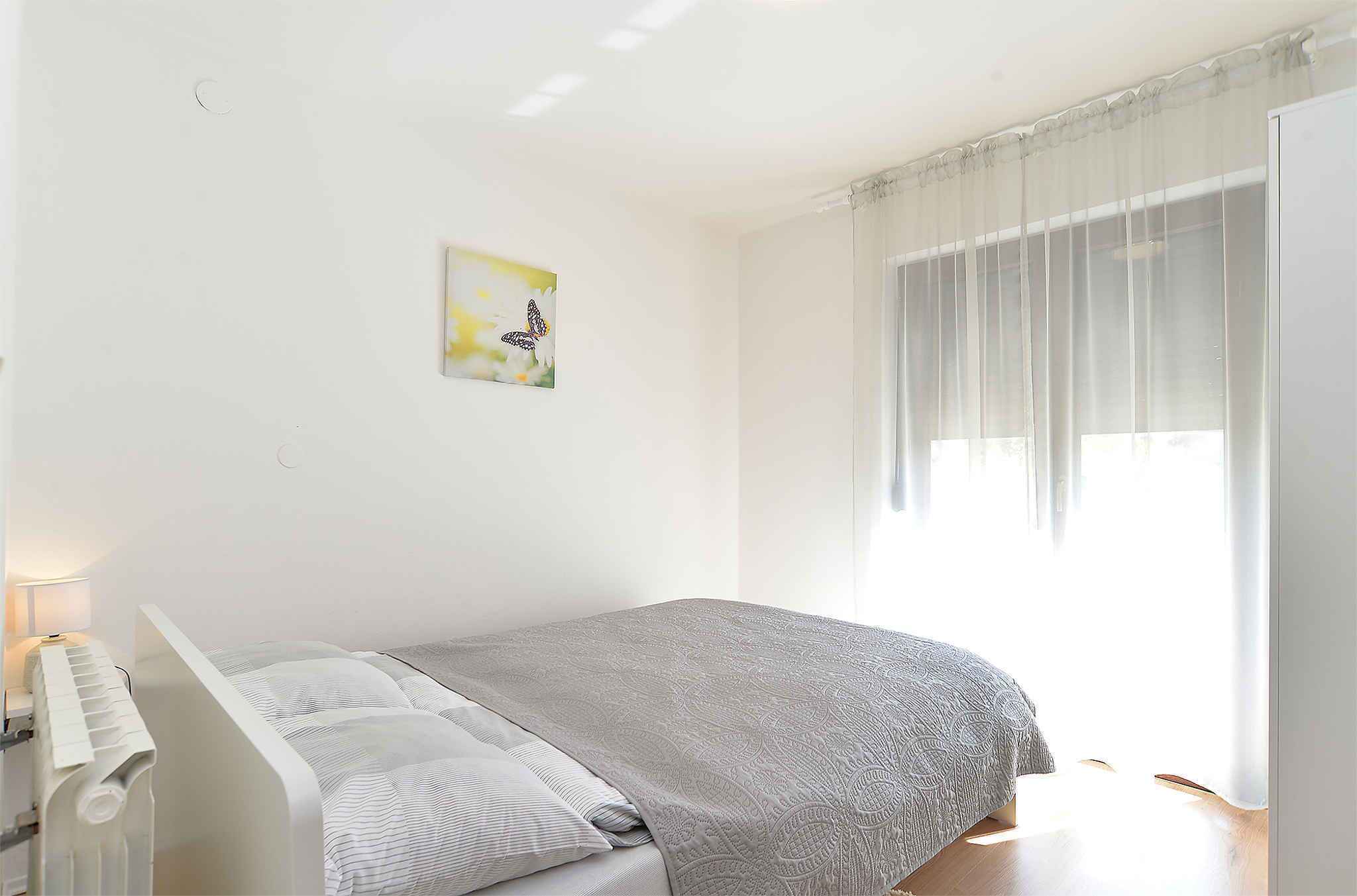 Un dormitorio con cama, radiador, ventana con cortinas translúcidas y un cuadro colgado en la pared.
