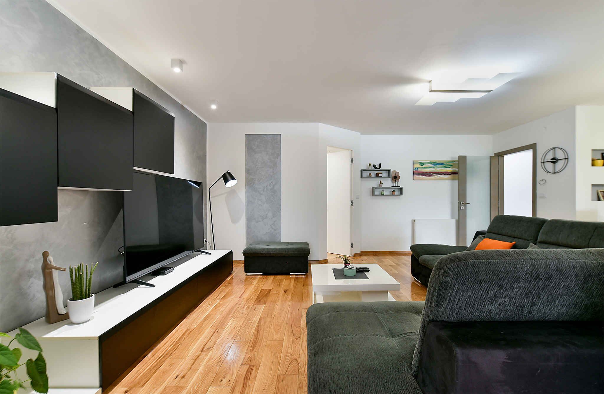Een moderne woonkamer met een flatscreen televisie, grijze banken, een salontafel en een minimalistisch design.