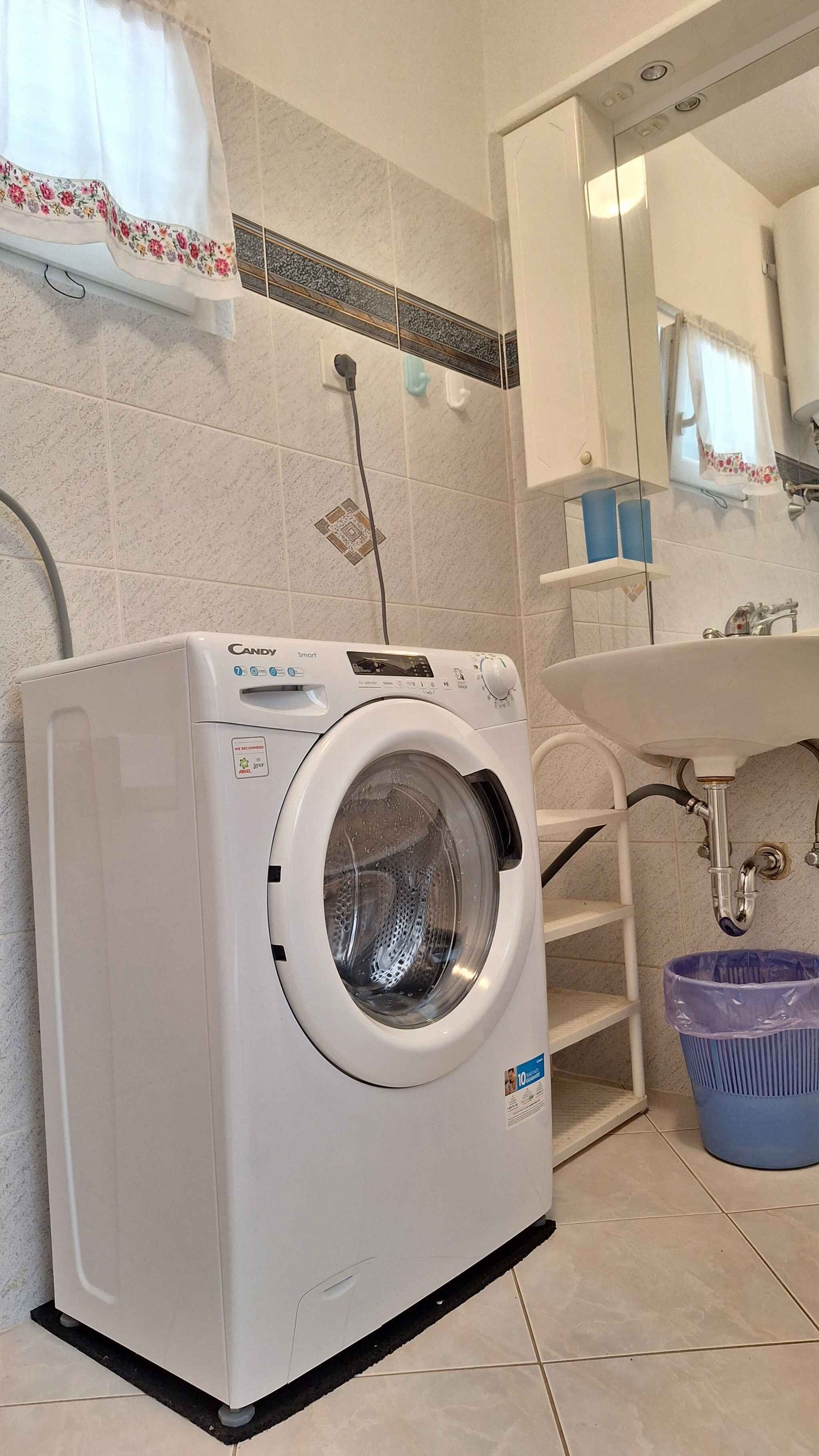 A white Candy Smart washing machine in a bathroom with a sink, mirror, and blue trash bin.