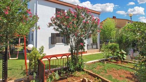 A white building with shutters, surrounded by a garden with various plants and a swing set.