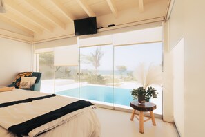 Un dormitorio con una cama grande, una silla, una mesita con una planta y un ventanal corredizo que da a una piscina.