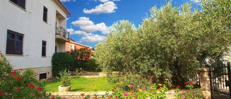 A white building with a balcony, surrounded by a garden with various plants and flowers.