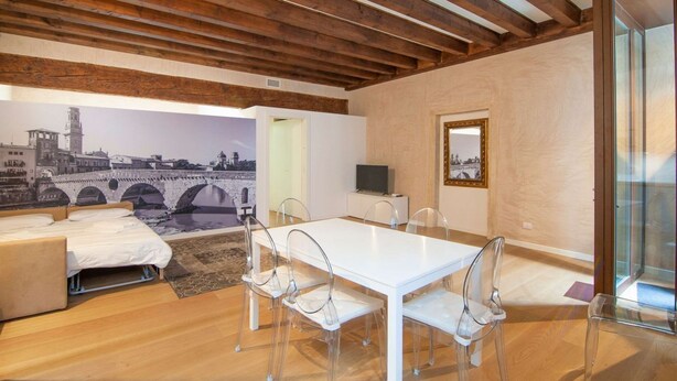 A modern room with a white dining table, clear chairs, a sofa, a TV, and a mural of a bridge.