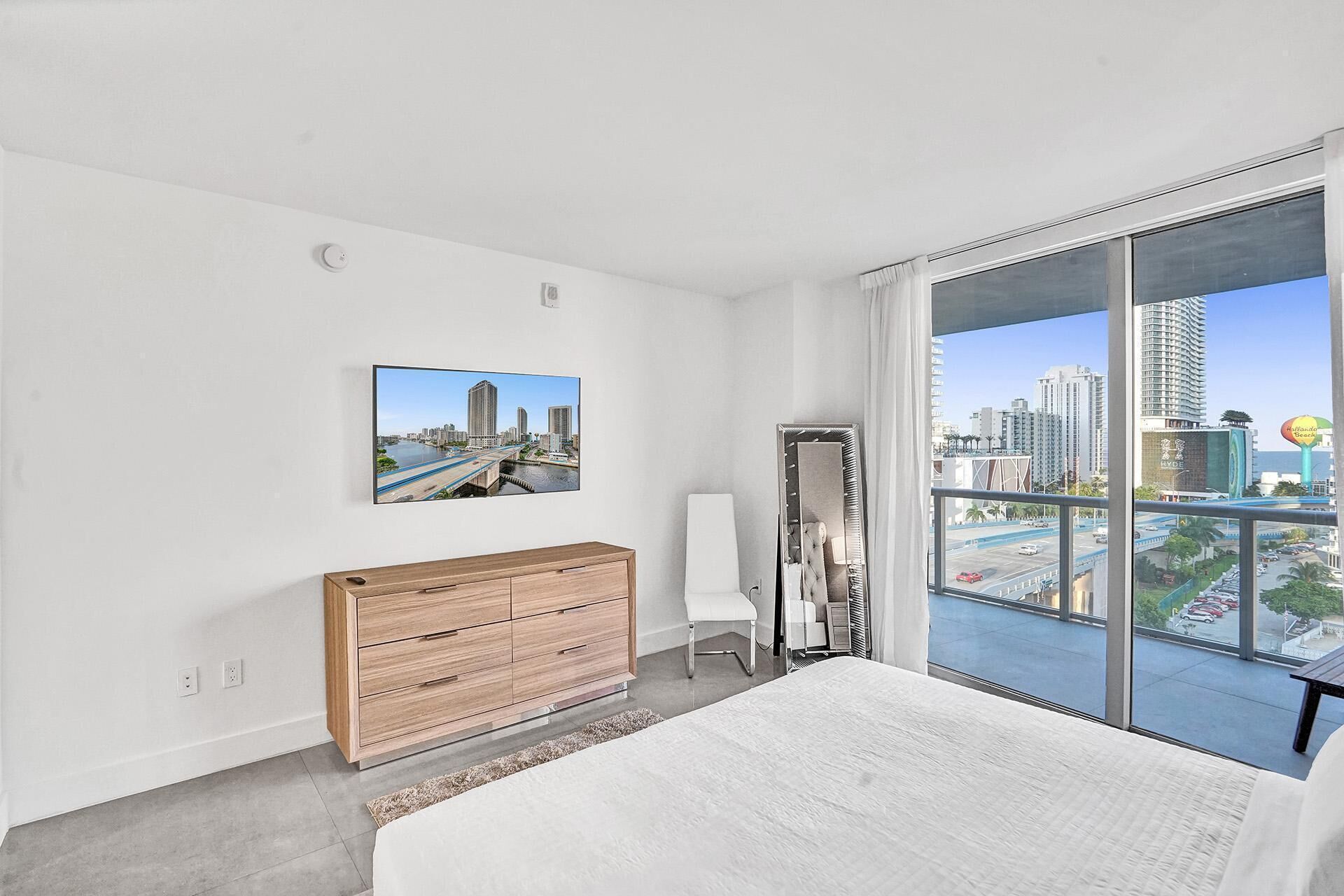 A modern bedroom with a large bed, a wooden dresser, a mirror, and a city view through large windows.