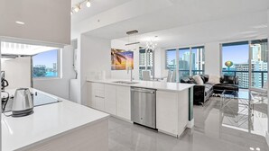 Eine moderne Küche mit Geschirrspüler, ein Essbereich mit einer schwarzen Couch und einem Glastisch sowie große Fenster mit Blick auf die Stadt.