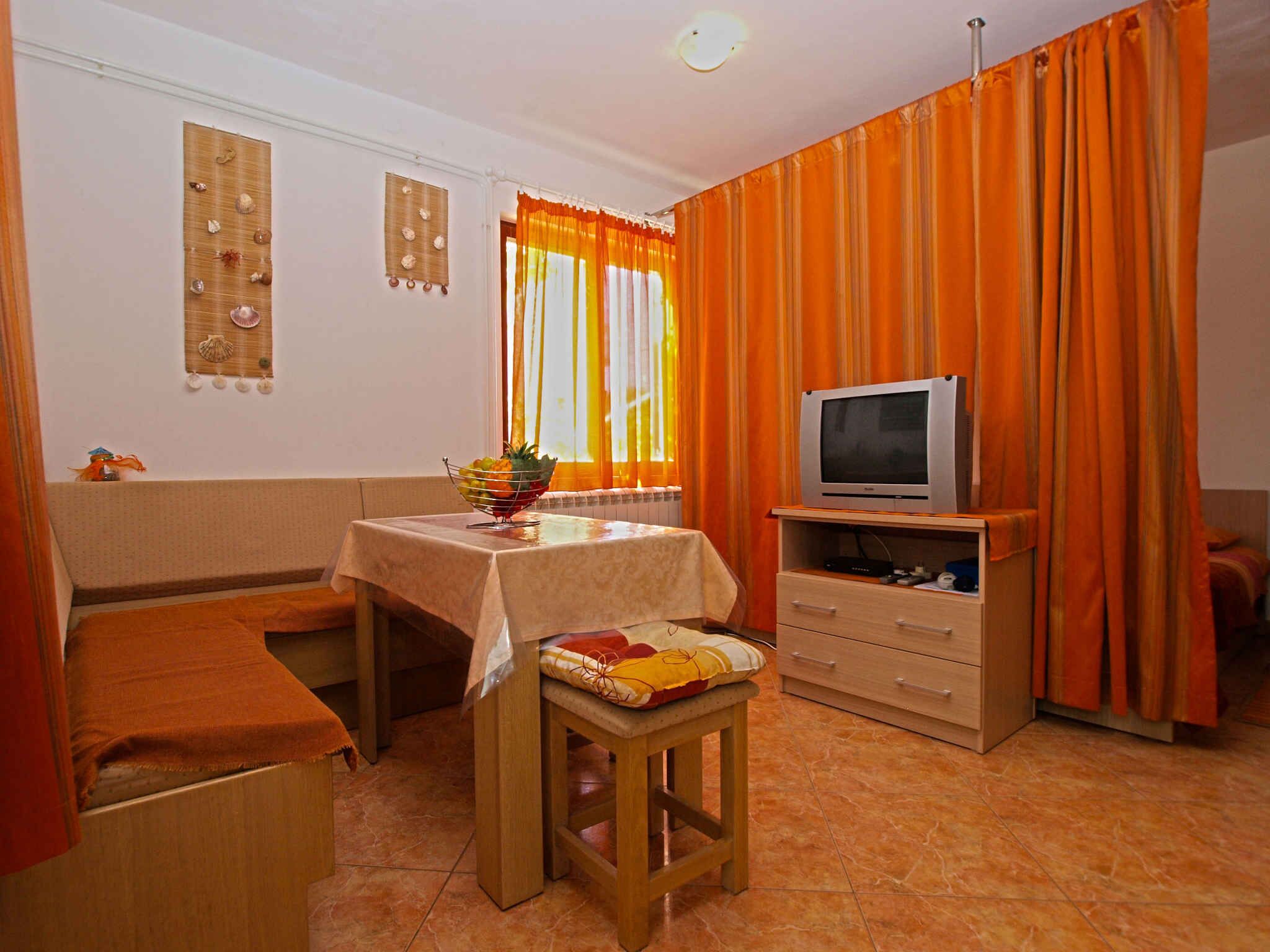 Une pièce de taille modeste, avec un lit, une petite table sur laquelle se trouve un saladier rempli de fruits, un téléviseur posé sur un meuble en bois et des rideaux orange.