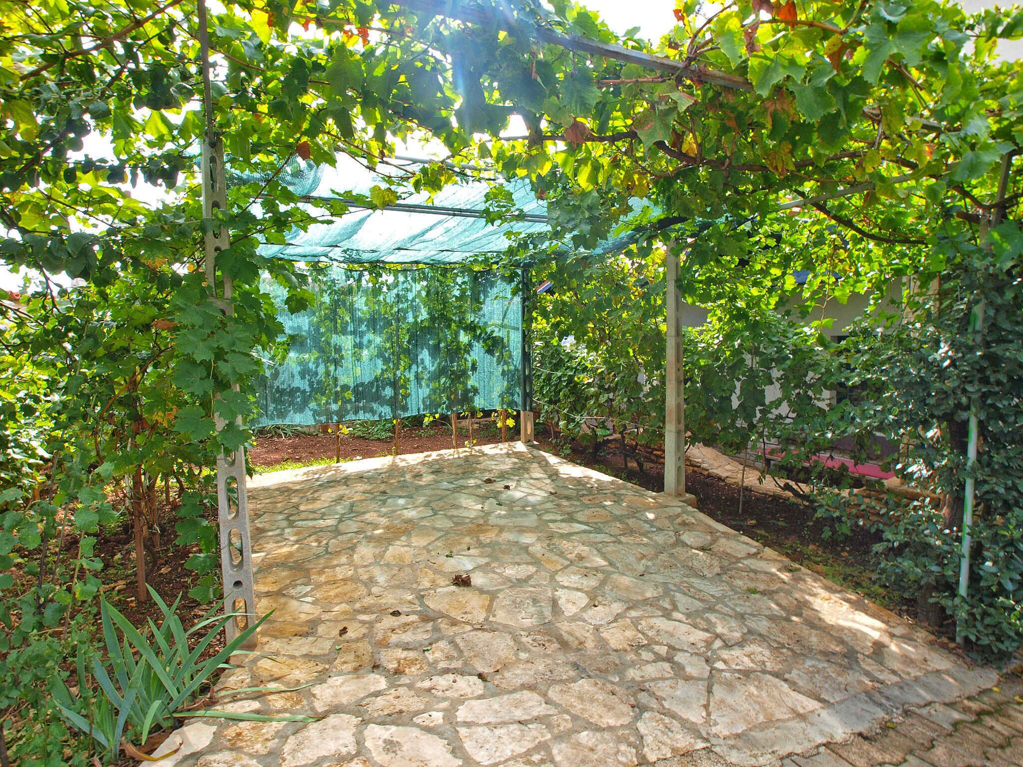 A stone-paved patio with a pergola covered by a green netting, surrounded by lush greenery.