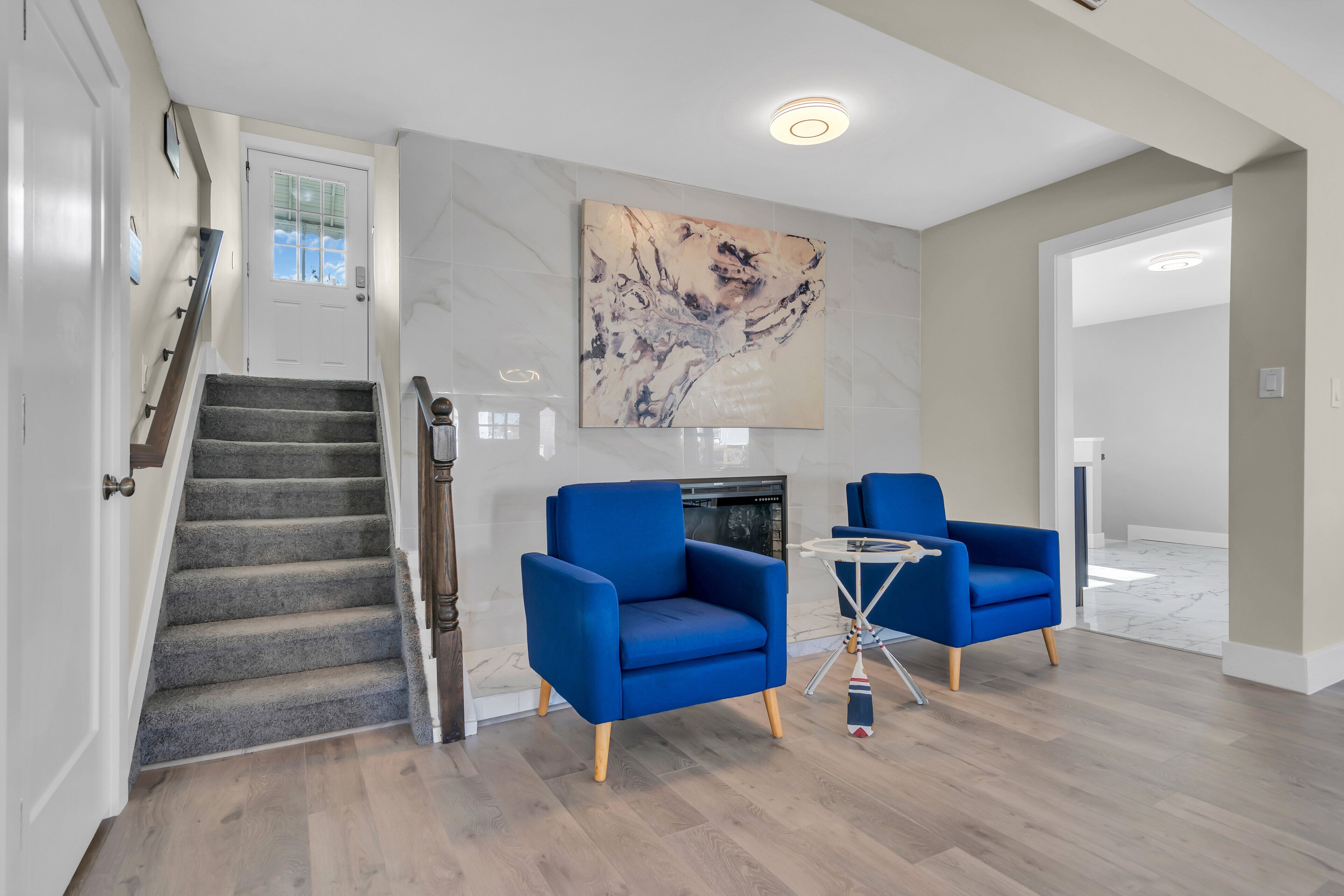 A modern living room with a fireplace, two blue armchairs, a small table, and a staircase.
