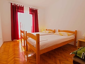 A room with a wooden bed, two pillows, a nightstand with a lamp, and a window with red curtains.