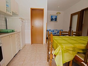 Una pequeña cocina con un refrigerador, un lavamanos y armarios. Se ve una puerta de madera que da a otra habitación. En primer plano, una mesa de comedor con sillas.