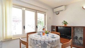 Un comedor con una mesa redonda cubierta por un mantel de encaje blanco, dos sillas, una planta en maceta, un televisor y un aparador con puertas de vidrio.