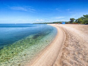A beach with clear shallow waters, a sandy shore, and a few buildings in the background.