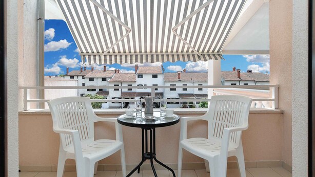 A balcony with a striped awning, two white plastic chairs, a small round table, and a view of neighboring buildings. - Holiday apartment with air conditioning (Pula)