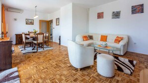 Un'ampia zona giorno con pavimento in parquet, un divano bianco con cuscini arancioni, un tavolino da caffè in vetro e una zona pranzo con mobili in legno.