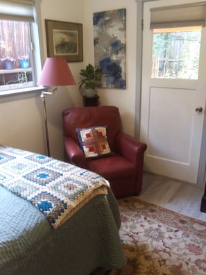 Une pièce comprenant un lit, un fauteuil rouge avec un coussin matelassé, une lampe sur pied, un tableau et une porte donnant sur l’extérieur.