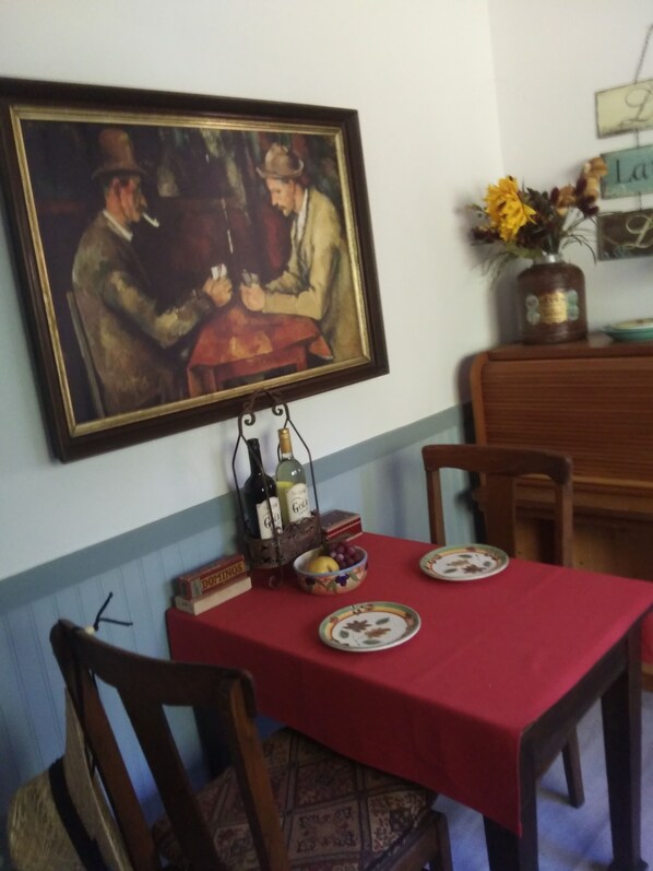 A dining area with a red tablecloth, two chairs, a painting, a vase with flowers, and a bottle holder with wine bottles.