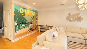 A living room with a beige sofa, a wooden floor, a wall mural of a van and surfboard, and a chandelier. - The Palmetto @ Park Circle - 10 Minutes From Downtown, The Palmetto @ Park Circle (North Charleston)
