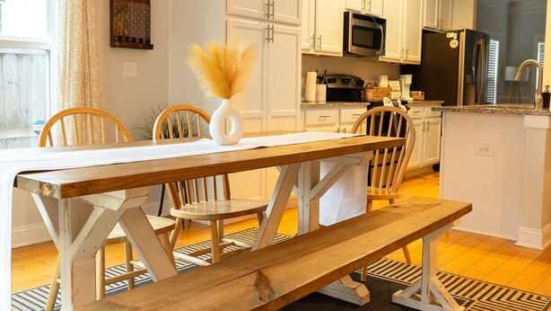 A modern kitchen with white cabinets, a wooden dining table with benches, and a central island with a microwave and refrigerator. - The Palmetto @ Park Circle - 10 Minutes From Downtown, The Palmetto @ Park Circle (North Charleston)