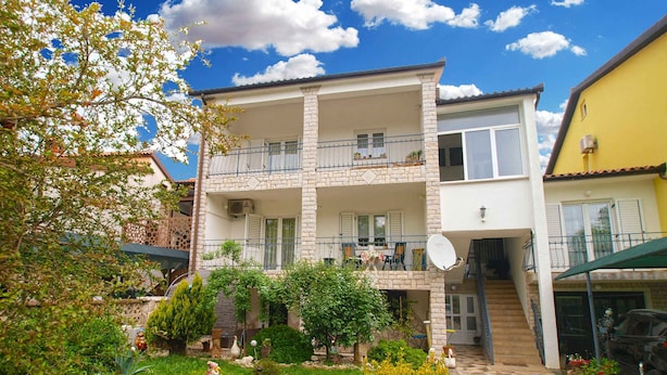 A two-story building with a balcony, a garden with various plants and a statue, and a clear blue sky.