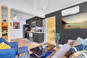 Un salon moderne avec un canapé, une table basse et un coin cuisine doté de meubles sombres et d’une porte jaune.