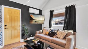 A living room with a sofa, a coffee table, a plant, a painting, and a window with curtains.