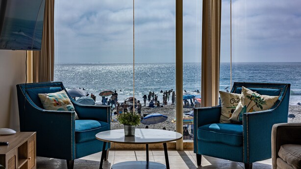 A room with a large window overlooking a beach, two blue armchairs, a small round table with a plant, and a flat-screen TV mounted on the wall.