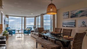 A spacious living area with a glass table, wicker furniture, and a large window overlooking the sea.