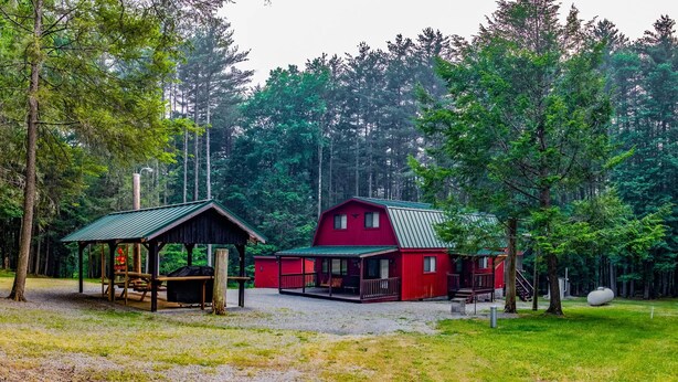 Une cabane rouge avec un porche et un petit abri, entourée d’une forêt.