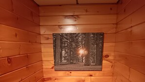 L’intérieur d’un chalet en bois, avec un tableau encadré représentant une scène de forêt accroché au mur, une fenêtre en bas et un escalier en bois.