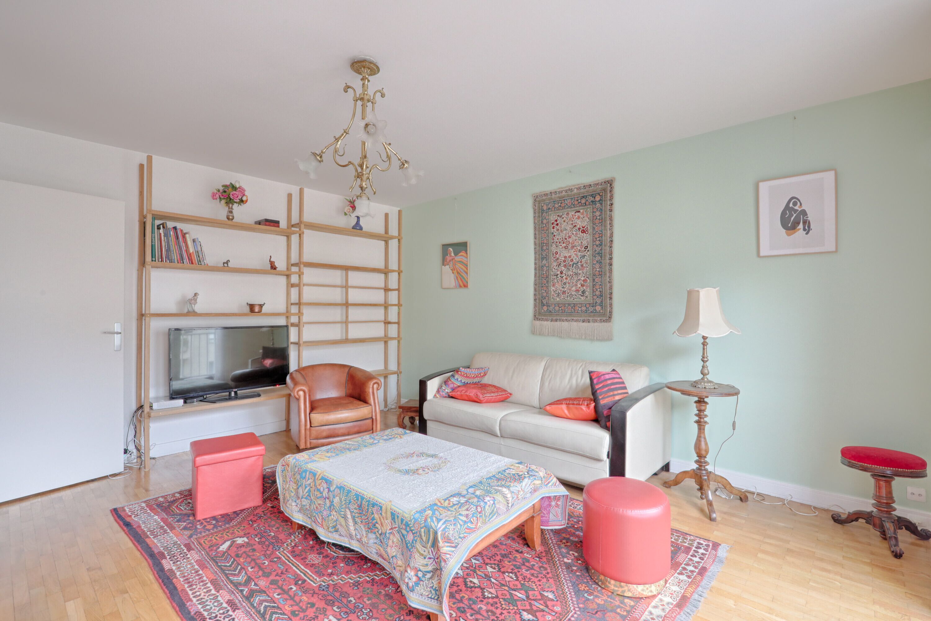 Ein Wohnzimmer mit Fernseher, Sofa, Pouf in Rot, Couchtisch, Bücherregal und einer Chandelier.