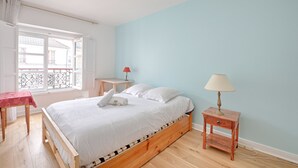 A bedroom with a wooden bed, a small bedside table with a lamp, a window with a view of buildings, and a table with a red tablecloth. - Appartement Martino - Welkeys (Paris)