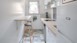 A compact kitchen with a window, a small stool, a kettle, and a refrigerator. - Appartement Martino - Welkeys (Paris)