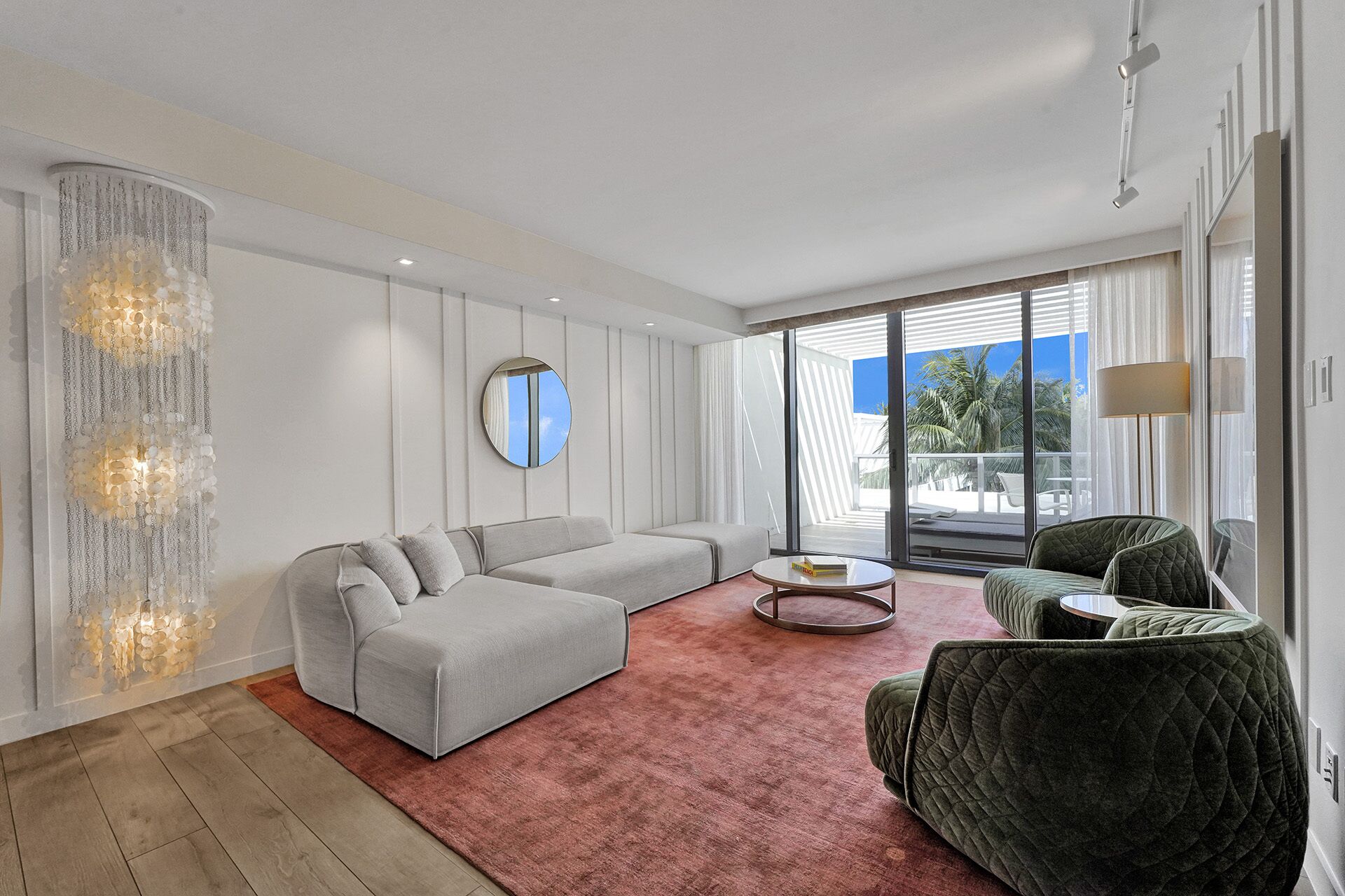 A modern living room with a grey sectional sofa, a round coffee table, and a dark green armchair. A large mirror hangs on the wall, and there is a view of the outdoors through large windows.