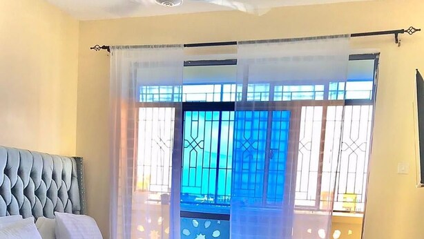 Un dormitorio con una cama grande, un cabecero tapizado en azul, un balcón con vistas y un ventanal con barandillas decorativas.