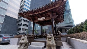 A traditional wooden pavilion with a sign, stone lion statues, and a modern building in the background.
