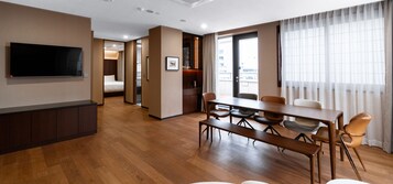 Una habitación de hotel moderna con suelo de madera, un área de comedor con mesa y sillas, un televisor montado en la pared y un ventanal con cortinas.